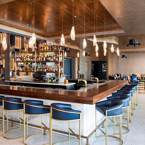 The image shows a modern bar with a wooden ceiling, hanging light fixtures, a fully stocked bar, blue bar stools, and seating in the background.