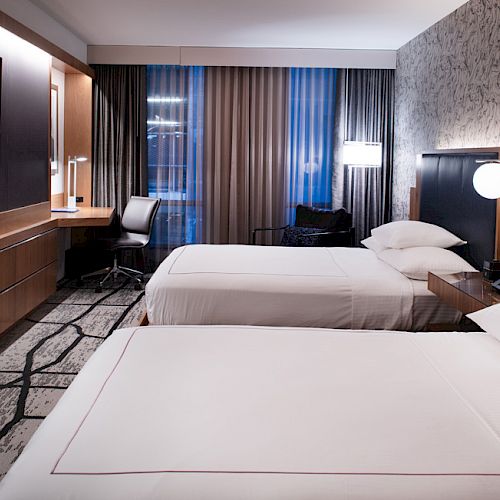 A modern hotel room with two beds, a flat-screen TV, a desk with a chair, ambient lighting, and floor-to-ceiling windows with curtains ending the sentence.
