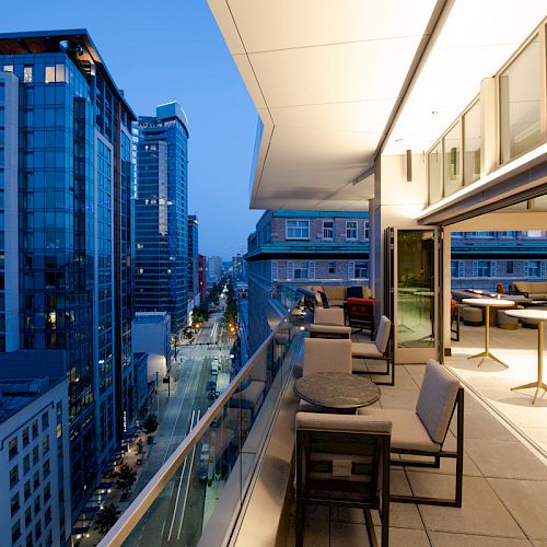 A modern urban balcony with seating overlooks a city street lined with high-rise buildings, illuminated in the evening.
