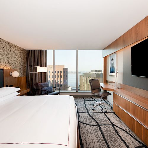 A modern hotel room features a large bed, desk, armchair, flat-screen TV, and a large window with an urban view.