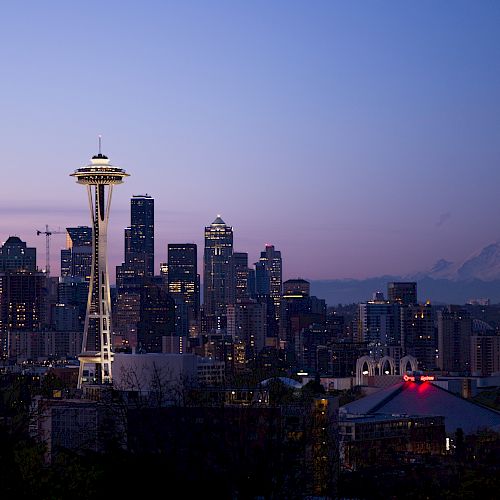 A city skyline at dusk features the Space Needle lit up, with tall buildings and a mountain in the background under a purple sky.