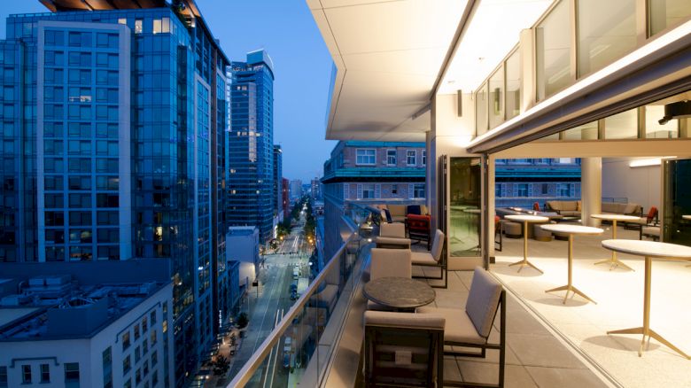 The image shows a modern cityscape from a high balcony with outdoor seating and tables, overlooking buildings and a street illuminated at twilight.