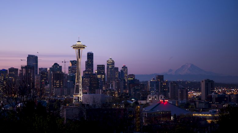 This image shows the Seattle skyline at dusk, featuring the Space Needle prominently lit up, with Mount Rainier in the background.