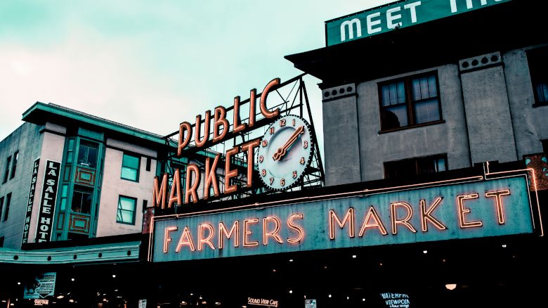 The image shows the iconic sign for a public farmers market, with a clock, situated among older buildings under a cloudy sky.