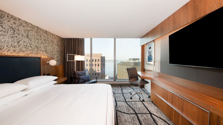 A modern hotel room with a double bed, large TV, desk, and a city view through a floor-to-ceiling window.