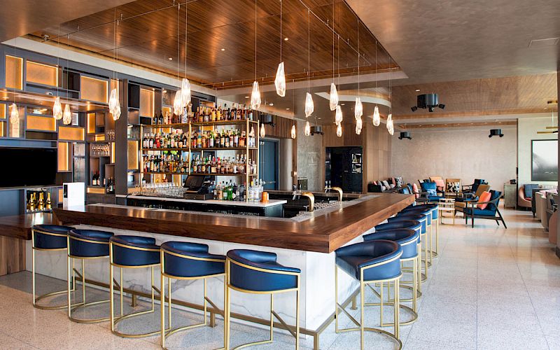 A modern bar interior with a central counter, blue stools, pendant lights, and a fully stocked bar. The area also has seating and a contemporary design.
