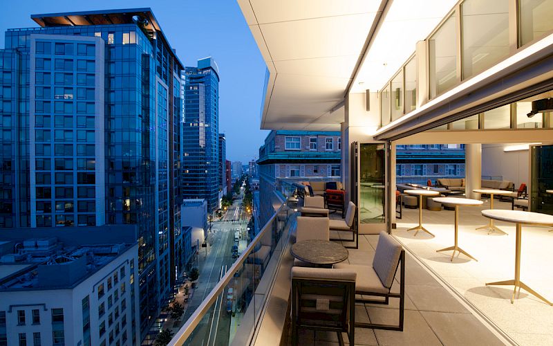 A modern balcony with furniture overlooks a bustling cityscape with high-rise buildings at dusk, featuring large windows and city lights.