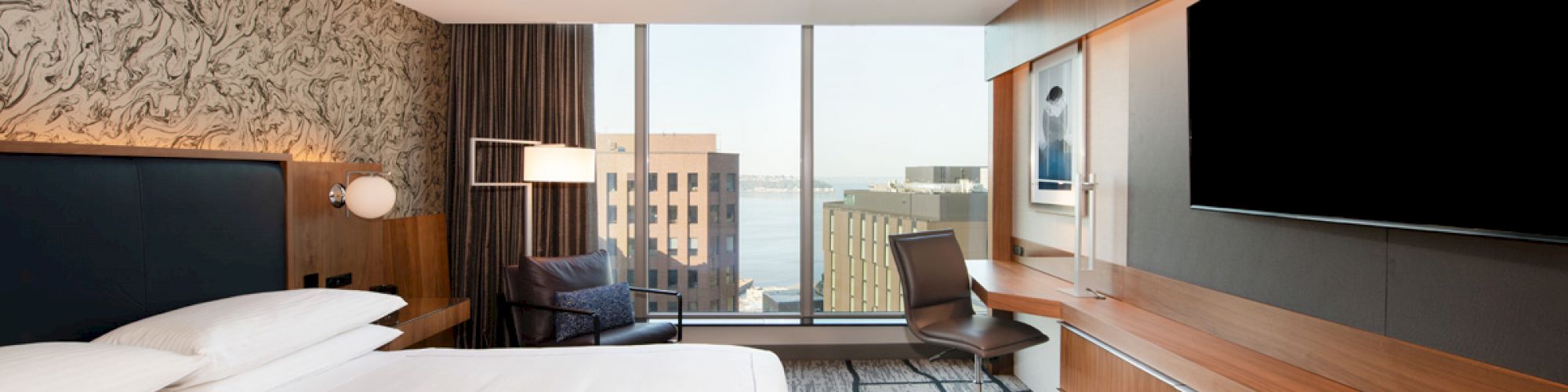 A modern hotel room with a bed, wall-mounted TV, desk, chair, and large windows offering a cityscape view.