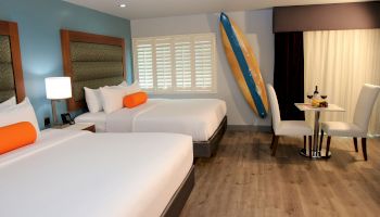 This image shows a hotel room with two beds, a surfboard, a small dining table, chairs, a bottle of wine, and a fruit plate, ending the sentence.