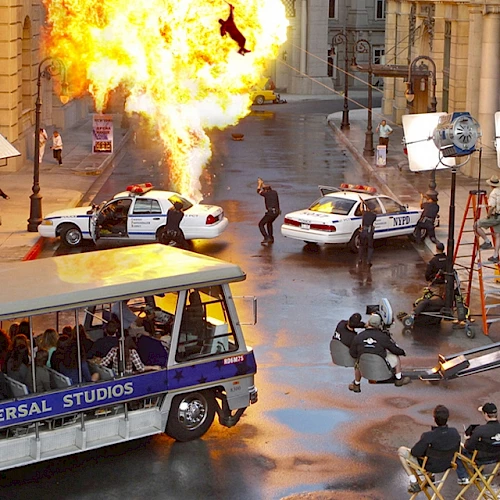 A film set with a staged explosion, police cars, and a tram labeled "Universal Studios" with passengers, surrounded by lights and crew members.